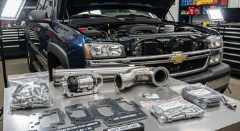 LBZ Duramax 6.6L engine bay with EGR delete kit components laid out on a workshop bench — block-off plates, stainless up-pipe, and intake elbow for 2006-2007 Chevrolet Silverado 2500HD